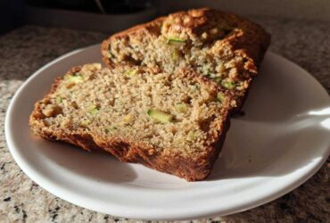 A close-up view of two slices of moist zucchini banana bread on a white plate, showing visible green zucchini pieces.