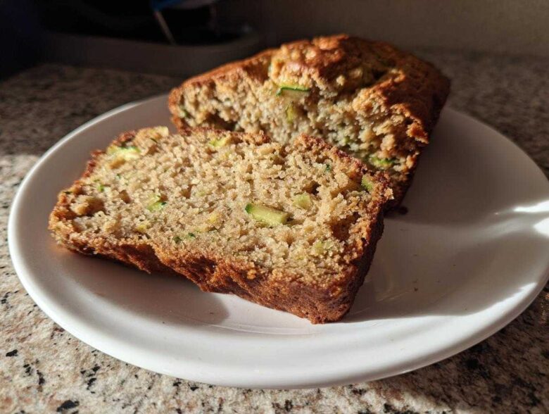 A close-up view of two slices of moist zucchini banana bread on a white plate, showing visible green zucchini pieces.