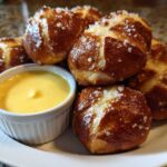 A plate of freshly baked Pretzel Bites sprinkled with coarse salt next to a small bowl of bright yellow beer cheese dip.