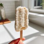 A hand holding one creamy Banana Coconut Popsicles coated in shredded coconut flakes.
