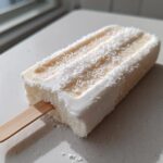 A single, creamy Banana Coconut Popsicles coated in shredded coconut resting on a light countertop.