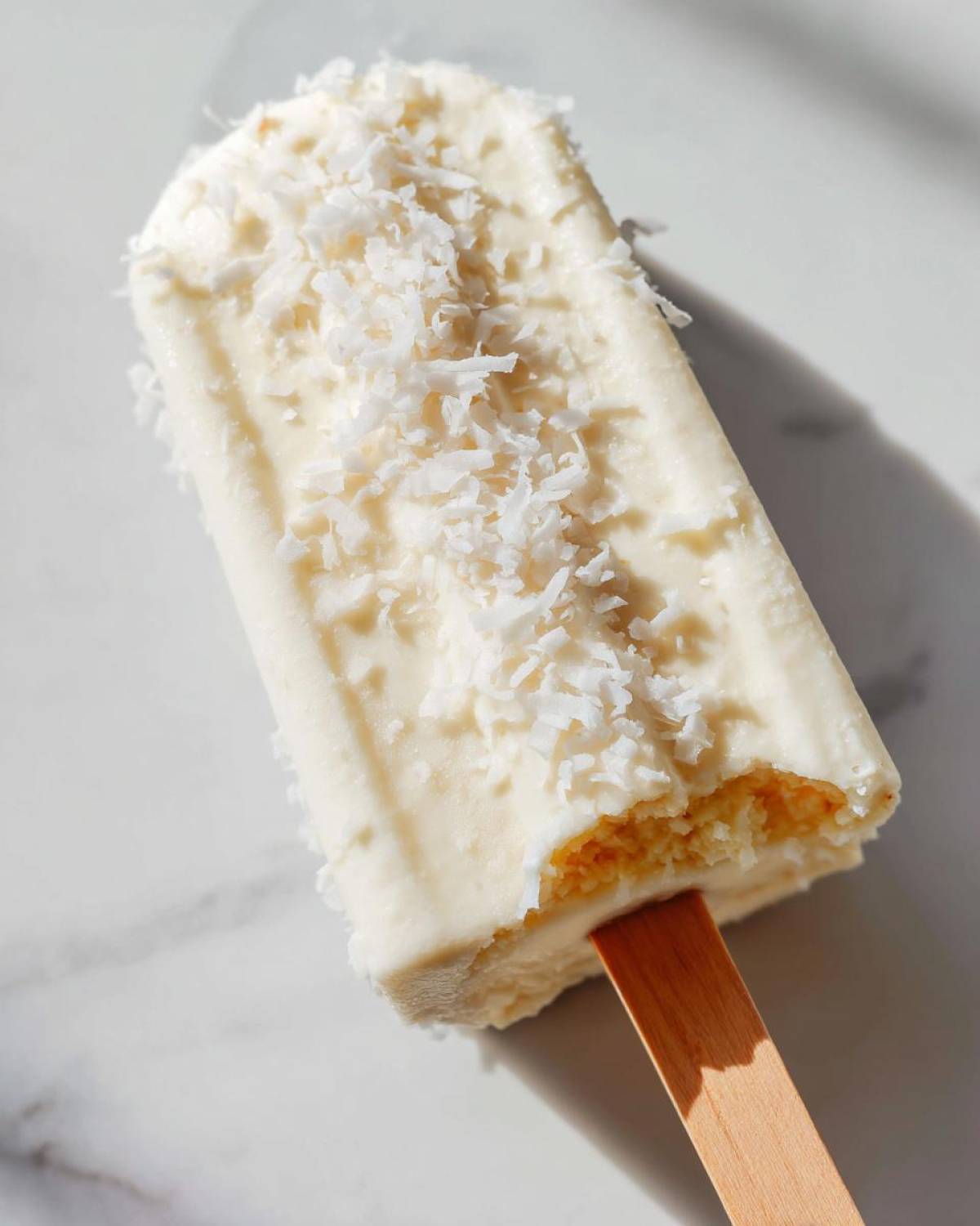 A creamy white Banana Coconut Popsicles topped with shredded coconut flakes, resting on a white marble surface.
