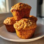 Four golden-brown Banana Oatmeal Muffins stacked on a white plate, catching warm morning sunlight.