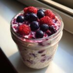 Close-up of creamy Overnight Oats topped generously with fresh raspberries and blueberries in a glass jar.