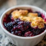 A close-up, appetizing view of warm Blackberry Cobbler bubbling with juicy berries and topped with golden biscuit crust in a white ramekin.