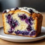 A close-up slice of rich Blueberry Cream Cheese Bread Loaf showing vibrant purple berries baked into the light yellow crumb and topped with white icing.