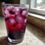 A tall, sweating glass filled with vibrant purple Blueberry Lemonade, ice cubes, and whole blueberries, sitting on a sunlit countertop.