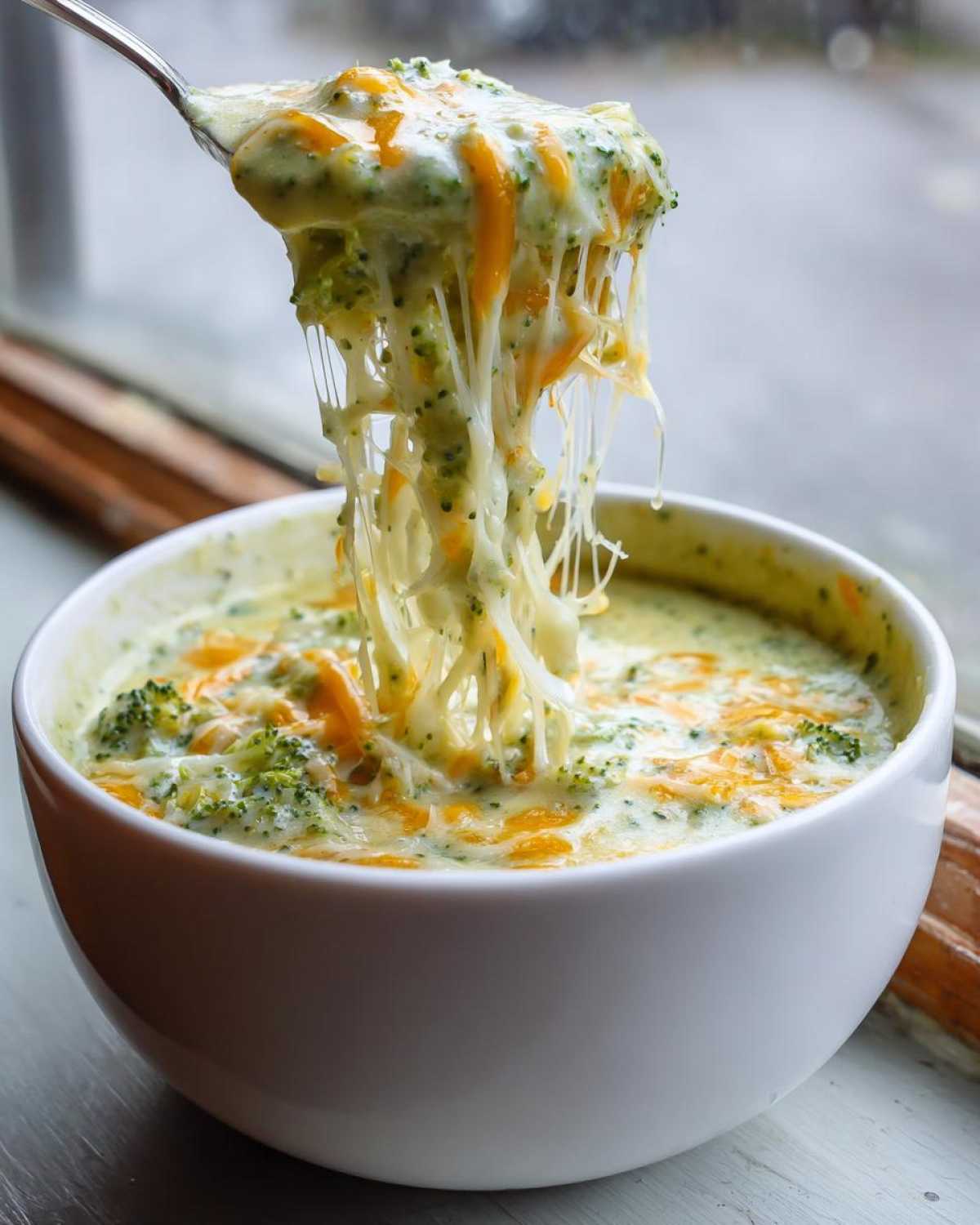 A spoonful of creamy Broccoli and Cheddar Soup showing extreme cheese pull over a white bowl.
