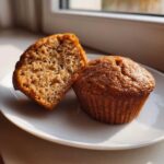 A close-up of two Brown Sugar Banana Muffins on a white plate, one sliced in half showing the moist interior crumb.