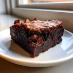 A single, rich square of Chewy Fudgy Brownies with a shiny top and visible chocolate chips, resting on a white plate.