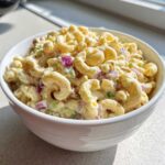 A close-up of Classic Homemade Macaroni Salad in a white bowl, featuring elbow macaroni coated in creamy dressing with visible chunks of red onion and celery.