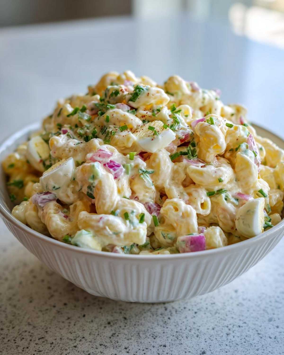 A close-up of a white bowl filled high with Classic Homemade Macaroni Salad, featuring elbow pasta, chopped eggs, and red onion.