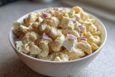 A close-up of a white bowl filled with creamy Classic Homemade Macaroni Salad containing elbow pasta, chopped red onion, celery, and hard-boiled egg.