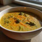 A close-up of a white bowl filled with creamy Broccoli and Cheddar Soup, topped with shredded cheese and broccoli florets.