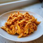 A close-up of shredded chicken coated in a rich, orange, creamy sauce, representing Creamy Crockpot Cajun Butter Chicken.