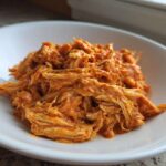 A close-up of shredded Creamy Crockpot Cajun Butter Chicken coated in a rich, orange sauce, served in a white bowl.