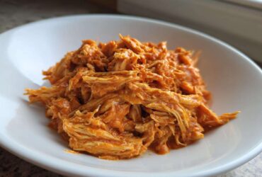 A close-up of shredded Creamy Crockpot Cajun Butter Chicken coated in a rich, orange sauce, served in a white bowl.