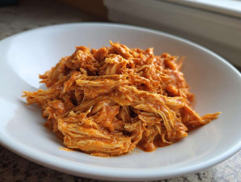 A close-up of shredded Creamy Crockpot Cajun Butter Chicken coated in a rich, orange sauce, served in a white bowl.