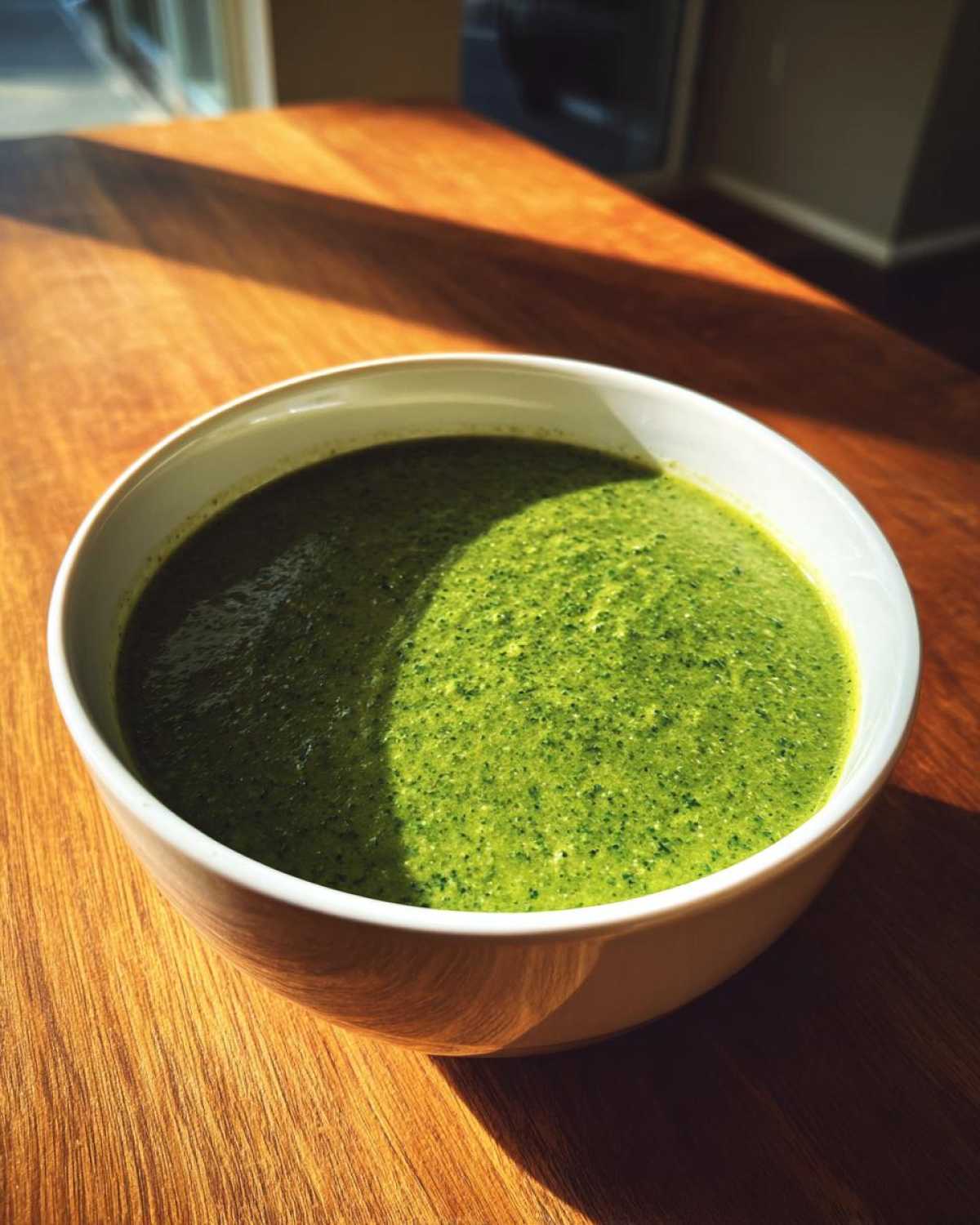A close-up of a white bowl filled with vibrant green Creamy Garlic Broccoli Soup, sitting on a wooden table bathed in sunlight.