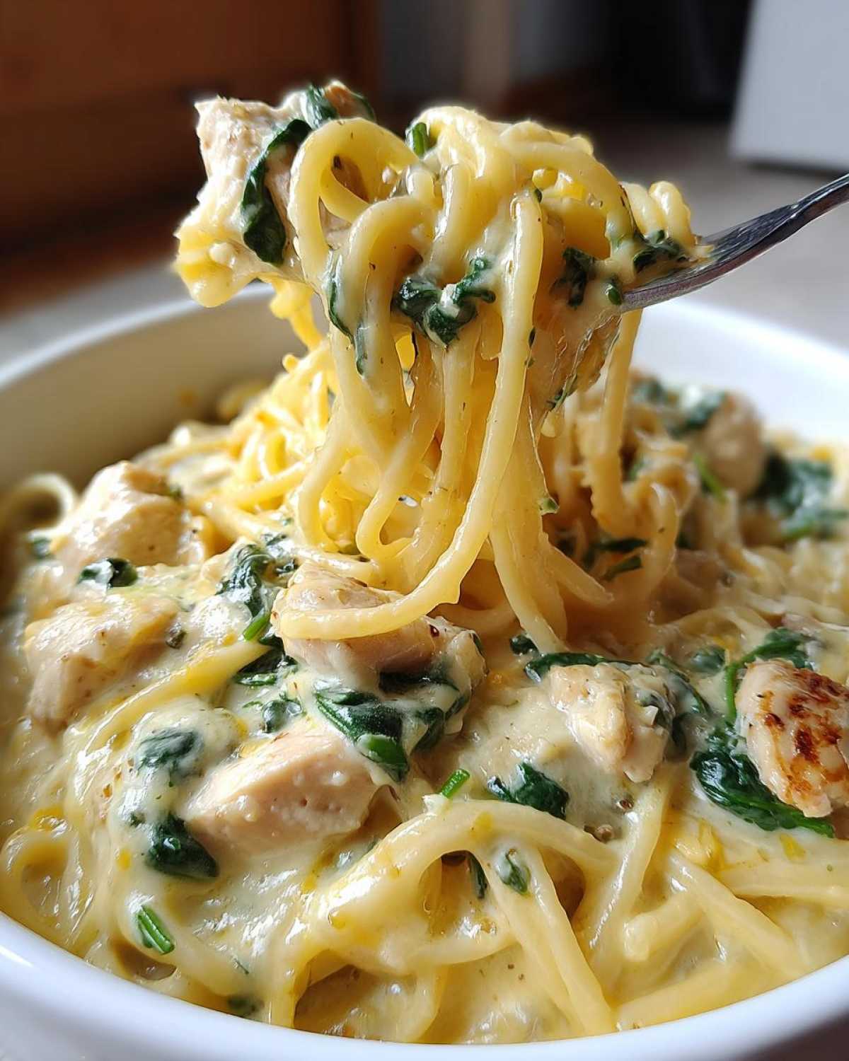 Close-up of creamy Lemon Chicken Pasta with spinach being lifted high from a white bowl on a fork.