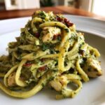 A close-up serving of creamy pesto pasta tossed with chunks of chicken and sun-dried tomatoes on a white plate.