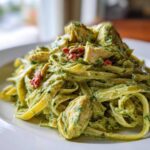 A close-up of creamy pesto pasta tossed with chunks of chicken and sun-dried tomatoes served on a white plate.