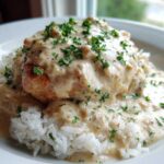 A close-up of a piece of chicken smothered in creamy sauce served over white rice, garnished with parsley, perfect for Creamy Smothered Chicken and Rice.