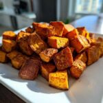 A mound of golden brown, cubed Crispy Roasted Sweet Potatoes seasoned with spices, served on a white rectangular plate.