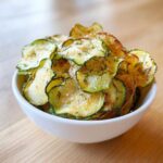 A white bowl filled with crispy, seasoned Crunchy Ranch Cucumber Chips sprinkled with visible herbs.