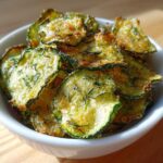A white bowl filled with golden brown, seasoned Crunchy Ranch Cucumber Chips, garnished with herbs.