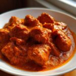 Close-up of tender chicken pieces swimming in a vibrant, orange-red sauce of Butter Chicken, served in a white bowl.