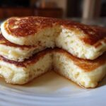 Close-up of a stack of three fluffy buttermilk pancakes, cut open to show the airy interior, drizzled with syrup.