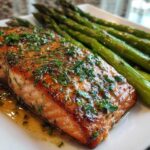 Close-up of a perfectly cooked fillet for a Garlic Butter Baked Salmon Dinner, glistening with sauce and topped with parsley, next to roasted asparagus.