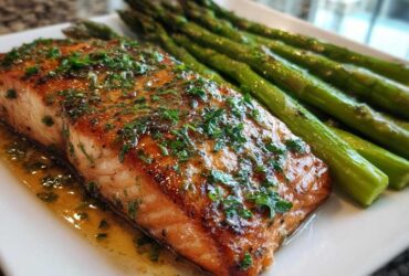 Close-up of a perfectly cooked fillet for a Garlic Butter Baked Salmon Dinner, glistening with sauce and topped with parsley, next to roasted asparagus.