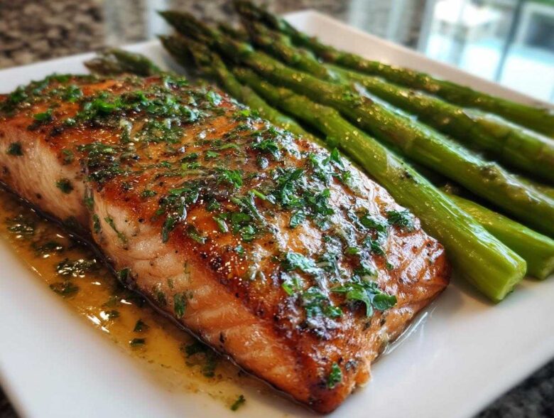 Close-up of a perfectly cooked fillet for a Garlic Butter Baked Salmon Dinner, glistening with sauce and topped with parsley, next to roasted asparagus.