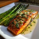 Close-up of a perfectly cooked Garlic Butter Baked Salmon Dinner fillet topped with minced garlic and herbs, served next to roasted asparagus.