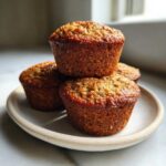 A stack of four golden-brown Banana Oatmeal Muffins resting on a small, light-colored ceramic plate.
