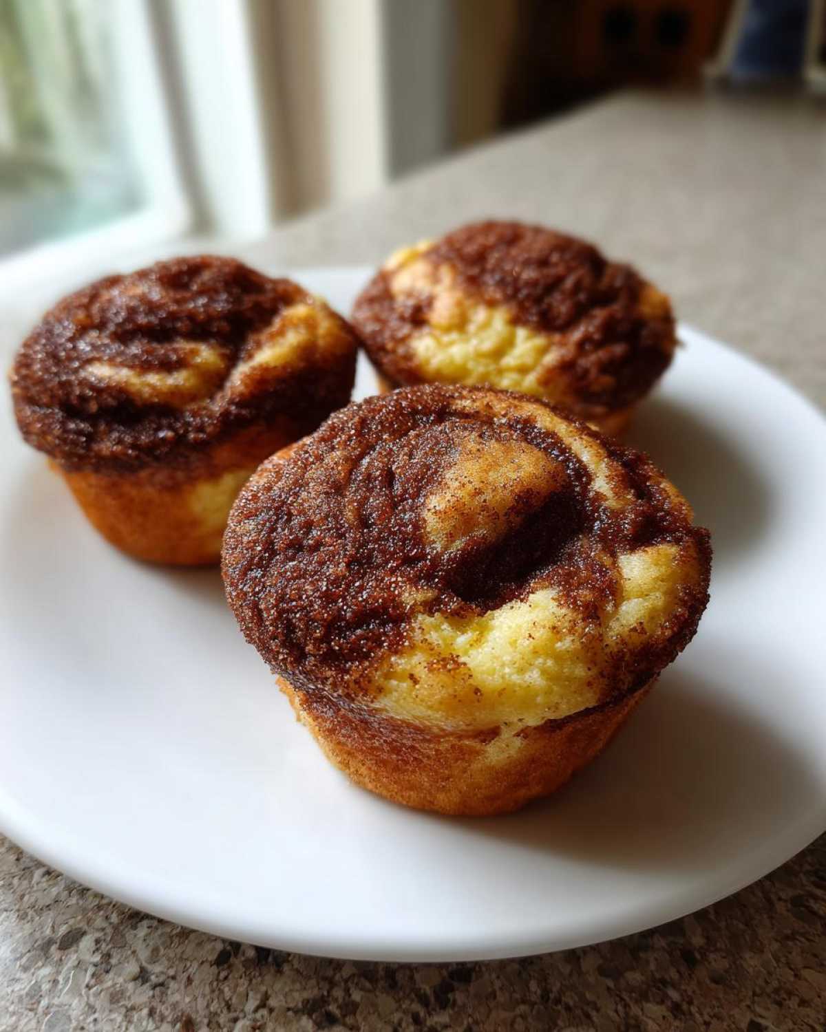 Three freshly baked Greek Yogurt Cinnamon Muffins with a dark cinnamon swirl topping, served on a white plate.