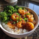 A white bowl filled with white rice topped with glossy Honey Garlic Shrimp, steamed broccoli, and garnished with sesame seeds and green onions.
