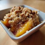 Close-up of a warm, individual serving of Peach Crumble with golden baked topping in a white square dish.