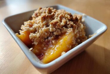 Close-up of a warm, individual serving of Peach Crumble with golden baked topping in a white square dish.
