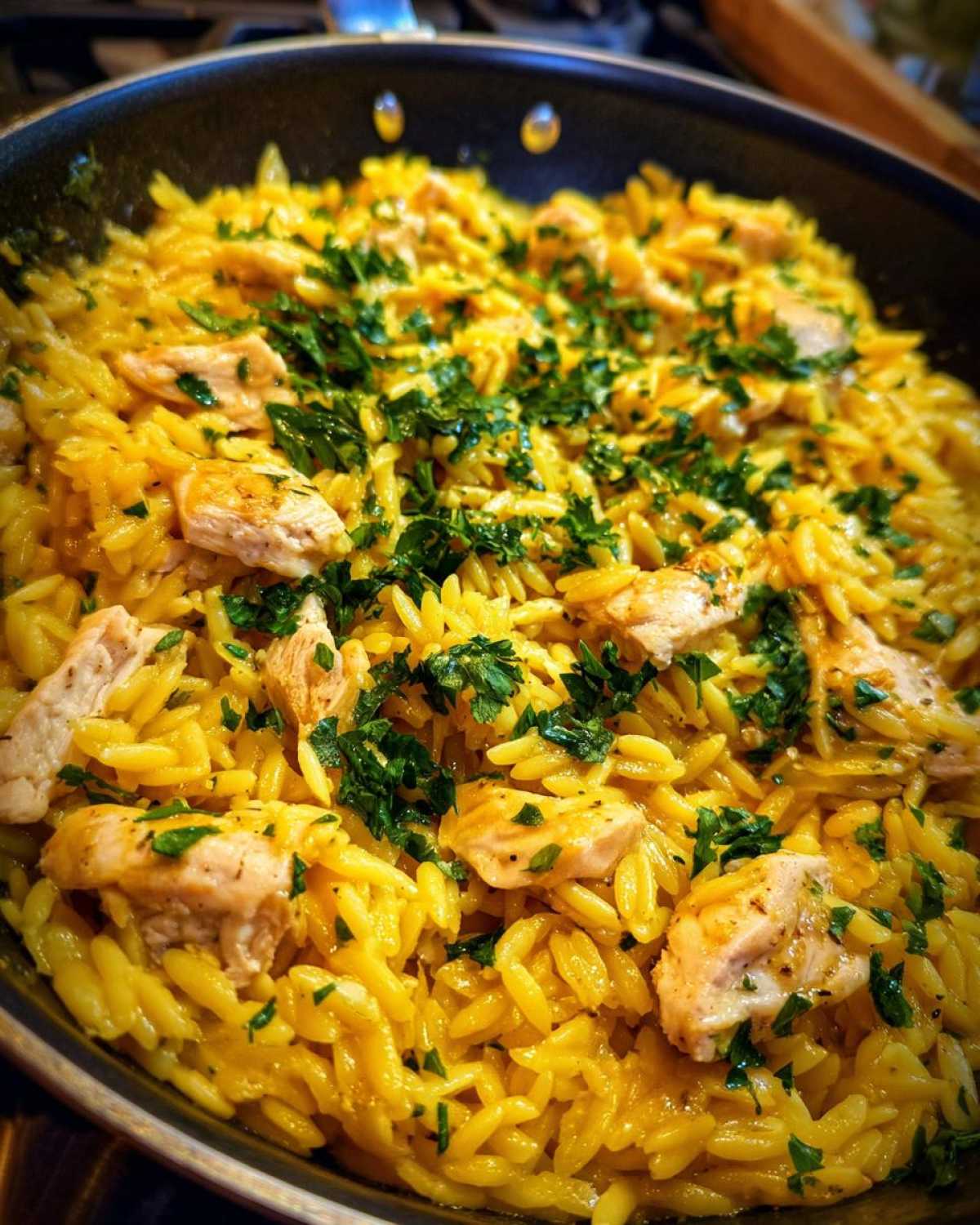 Close-up of bright yellow Lemon Chicken and Orzo dish garnished with fresh parsley in a black skillet.