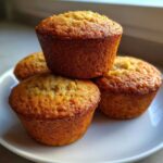 A close-up of four golden brown, perfectly baked Banana Muffins stacked slightly on a white plate.