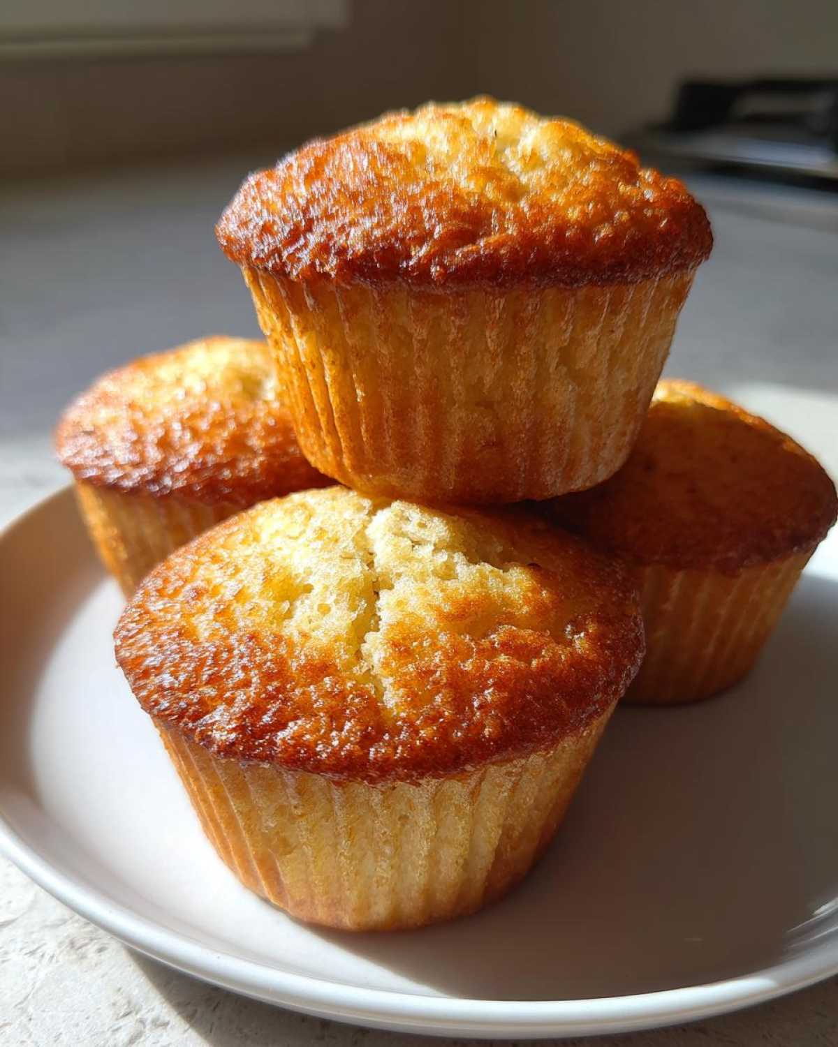 A stack of four perfectly baked, golden brown Banana Muffins resting on a white plate.