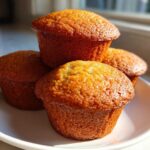 A stack of four perfectly baked, golden brown Banana Muffins sitting on a small white plate in natural sunlight.