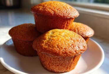 A stack of four perfectly baked, golden brown Banana Muffins sitting on a small white plate in natural sunlight.
