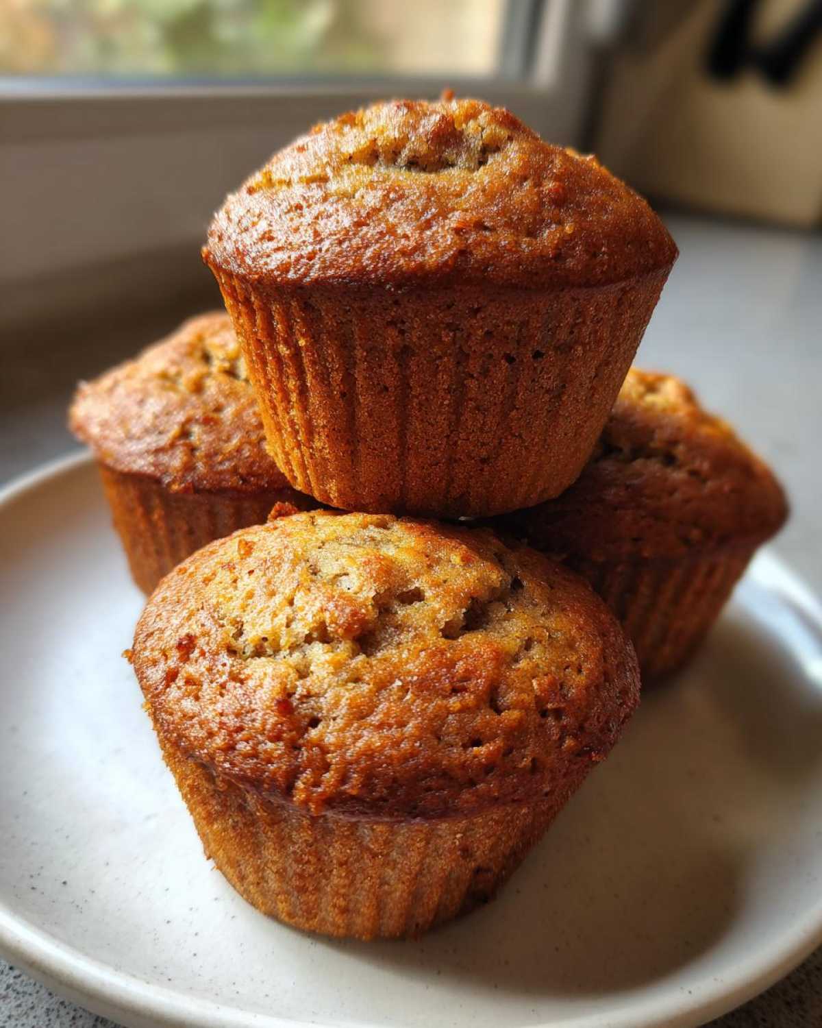 A stack of four golden-brown, moist Banana Muffins resting on a light-colored plate.
