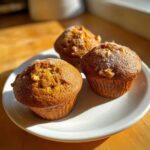 Three golden brown Banana Nut Muffins dusted with powdered sugar, served on a white plate.