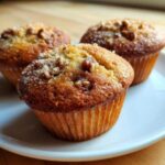 Three golden brown Banana Nut Muffins topped with chopped nuts and coarse sugar, resting on a white plate.