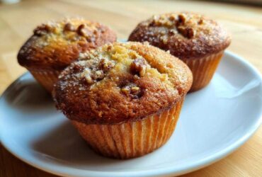 Three golden brown Banana Nut Muffins topped with chopped nuts and coarse sugar, resting on a white plate.